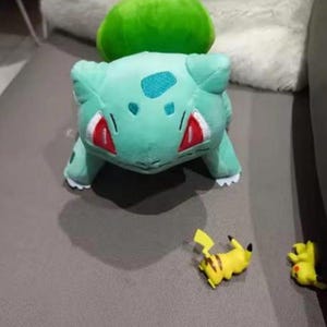 K&ouml;nnte beinhalten: Ein gr&uuml;nes Bulbasaur-Pl&uuml;schtier mit roten Akzenten und einer gr&uuml;nen Knolle auf dem R&uuml;cken. Zwei kleine gelbe Pikachu-Figuren sind ebenfalls vorhanden, was eine spielerische Szene ergibt.