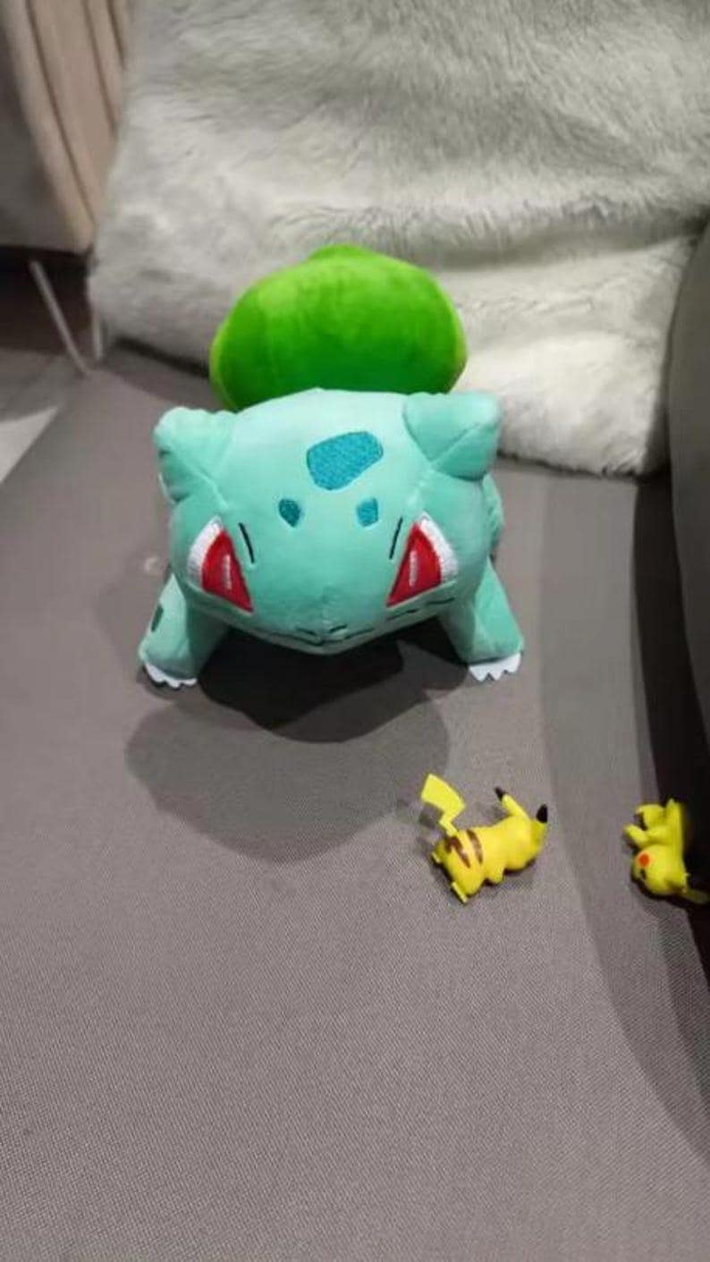 K&ouml;nnte beinhalten: Ein gr&uuml;nes Bulbasaur-Pl&uuml;schtier mit roten Akzenten und einer gr&uuml;nen Knolle auf dem R&uuml;cken. Zwei kleine gelbe Pikachu-Figuren sind ebenfalls vorhanden, was eine spielerische Szene ergibt.