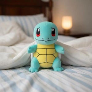 K&ouml;nnte beinhalten: Ein Stofftier von Squirtle, mit hellblauem K&ouml;rper, gelbem Panzer und roten Augen. Das Kuscheltier liegt auf einem Bett mit blau-wei&szlig; gestreiften Bettlaken und einer wei&szlig;en Decke.