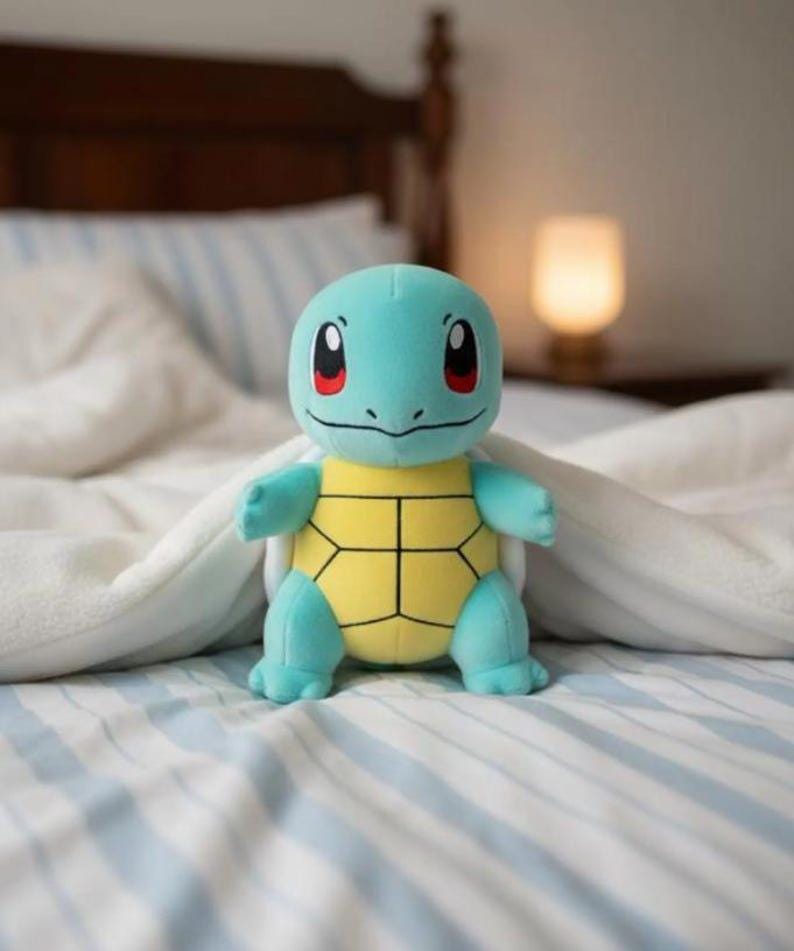 K&ouml;nnte beinhalten: Ein Stofftier von Squirtle, mit hellblauem K&ouml;rper, gelbem Panzer und roten Augen. Das Kuscheltier liegt auf einem Bett mit blau-wei&szlig; gestreiften Bettlaken und einer wei&szlig;en Decke.