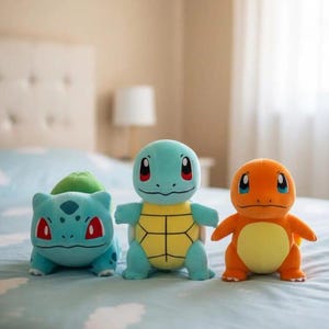K&ouml;nnte beinhalten: Drei Pok&eacute;mon-Pl&uuml;schfiguren: Bisasam, Schiggy und Glumanda. Bisasam ist t&uuml;rkis mit einer gr&uuml;nen Knolle, Schiggy ist blau mit einem gelben Panzer und Glumanda ist orange mit einem gelben Bauch. Sie liegen auf einer hellblauen Bettdecke.