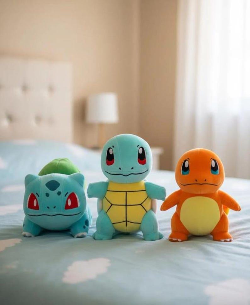 K&ouml;nnte beinhalten: Drei Pok&eacute;mon-Pl&uuml;schfiguren: Bisasam, Schiggy und Glumanda. Bisasam ist t&uuml;rkis mit einer gr&uuml;nen Knolle, Schiggy ist blau mit einem gelben Panzer und Glumanda ist orange mit einem gelben Bauch. Sie liegen auf einer hellblauen Bettdecke.