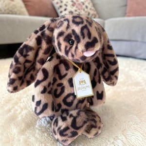 May include: A soft toy bunny featuring a leopard print design in brown and black. The bunny has long, drooping ears, a pink nose, and a tag that says "Jellycat". It is positioned on a white, fluffy surface.