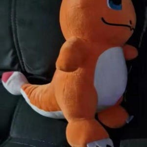 K&ouml;nnte beinhalten: Ein orangefarbenes Charmander-Pl&uuml;schtier mit wei&szlig;em Bauch und blauen Augen. Das Spielzeug hat einen langen Schwanz mit wei&szlig;er Spitze und sitzt auf einer dunklen Oberfl&auml;che.