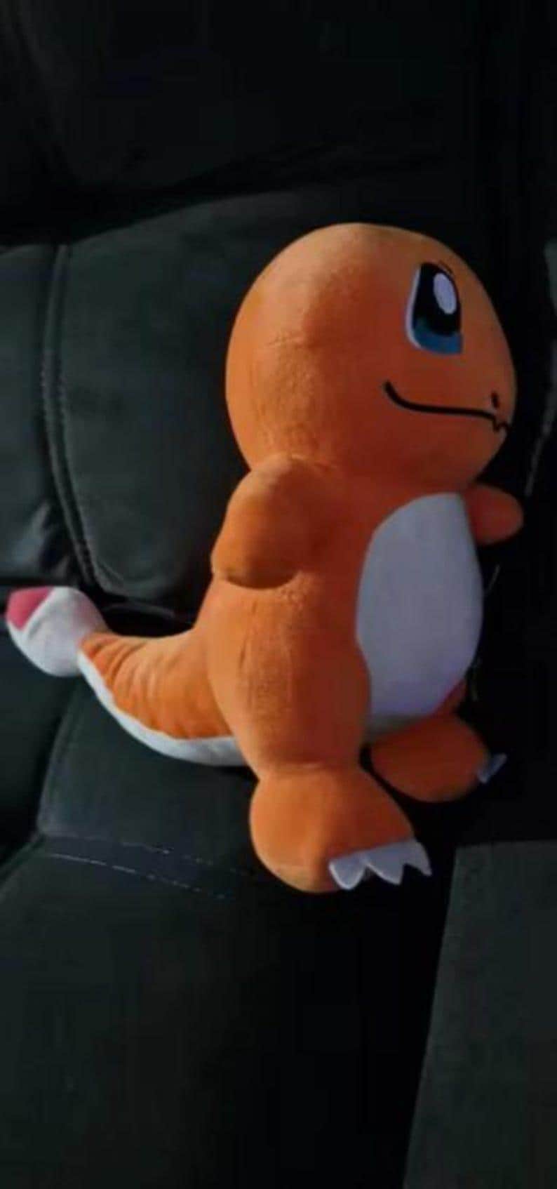 K&ouml;nnte beinhalten: Ein orangefarbenes Charmander-Pl&uuml;schtier mit wei&szlig;em Bauch und blauen Augen. Das Spielzeug hat einen langen Schwanz mit wei&szlig;er Spitze und sitzt auf einer dunklen Oberfl&auml;che.