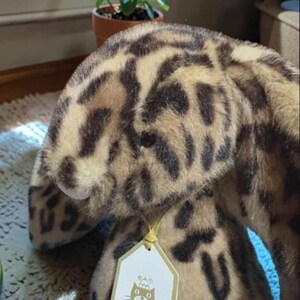 May include: A soft toy cat featuring a brown and black spotted design. The toy has a tag attached with a gold string. The cat is sitting on a white rug.
