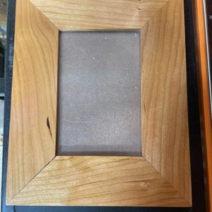 May include: A rectangular wooden picture frame with a light brown finish. The frame has a simple, classic design with a beveled edge. The frame is empty, with a gray backing visible inside.