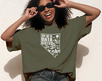 Op de afbeelding: Een olijfgroen T-shirt met korte mouwen en een witte grafische print in de vorm van de staat Nevada. Het ontwerp bevat de tekst "SAY NO TO DATA CENTERS" naast illustraties van bergen, een zon, een Joshua-boom en een biologisch gevarensymbool.