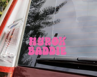 Op de afbeelding: Een roze vinyl sticker met de tekst "HYROK BADDIE" in een afgerond lettertype is op een autoruit geplakt. De sticker is te zien tegen het donkere glas, met ook het achterlicht van de auto en een ruitenwisser.