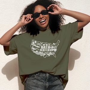 May include: An olive green t-shirt featuring a white graphic of the United States map filled with landscape illustrations and the text "SAY NO TO DATA CENTERS" in the center. The design includes mountains, trees, a farm, and a flag.