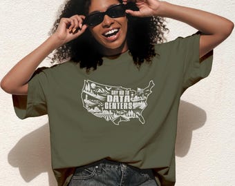 Op de afbeelding: Een olijfgroen t-shirt met een witte afbeelding van de kaart van de Verenigde Staten, gevuld met landschapsillustraties en de tekst "SAY NO TO DATA CENTERS" in het midden. Het ontwerp bevat bergen, bomen, een boerderij en een vlag.