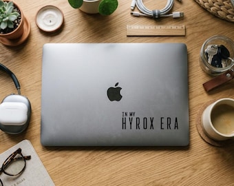Op de afbeelding: Een zilveren laptop met het Apple-logo en de tekst "IN MY HYROX ERA" op een houten bureau. Op het bureau staan ook een vetplant in een terracotta pot, een kaars, een koptelefoon, een notitieboek en een kopje koffie.