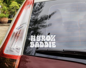 Op de afbeelding: Een witte vinyl sticker op een autoruit met de woorden "HYROX BADDIE" in een retro lettertype. De auto is donkerrood van kleur en het achterlicht is zichtbaar aan de linkerkant van de afbeelding.