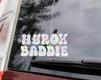 Op de afbeelding: Een autoraam met een holografische sticker met de tekst "HYROX BADDIE" in een bubbellettertype. De sticker heeft een regenboogeffect en is aangebracht op de achterruit van een bordeauxrode auto. De ruitenwisser is onderaan zichtbaar.