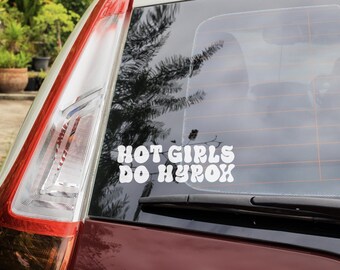 Op de afbeelding: Een witte vinyl sticker op een autoruit met de tekst "HOT GIRLS DO HYROX" in een retro lettertype. Het achterlicht van de auto is links zichtbaar, met een rood en wit ontwerp. De achtergrond toont bomen en een bewolkte lucht.
