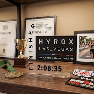 Peut inclure: Un trophée de finition en acrylique transparent avec le texte "HYROX LAS_VEGAS" et un temps de 2:08:35. Le trophée est exposé sur une étagère en bois avec d'autres souvenirs sportifs.