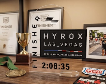 Op de afbeelding: Een heldere acryl finisher award met de tekst "HYROX LAS_VEGAS" en een tijd van 2:08:35. De award staat op een houten plank met andere sportmemorabilia.