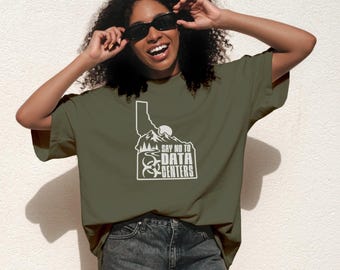 Op de afbeelding: Een olijfgroen T-shirt met korte mouwen en een witte afbeelding van de staat Idaho, bergen, een zon en een biohazard-symbool. De tekst "SAY NO TO DATA CENTERS" staat in een vetgedrukt bloklettertype in de omtrek van de staat.