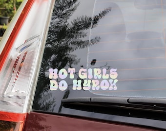 Op de afbeelding: Een autosticker met de tekst "HOT GIRLS DO HYROX" in een pastelkleurige regenbooglettertype. De sticker is aangebracht op de achterruit van een bordeauxrode auto, met het achterlicht zichtbaar aan de linkerkant.