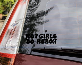 Op de afbeelding: Zwarte vinyl sticker met de tekst "HOT GIRLS DO HUROX" in een vetgedrukte retro lettertype, aangebracht op een autoruit. De afbeelding toont een deel van de rode carrosserie en het achterlicht van de auto, met een vage buitenachtergrond.