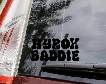 Op de afbeelding: Zwarte vinyl sticker met de woorden "HYROX BADDIE" in een retro lettertype, aangebracht op een autoruit. De sticker is te zien tegen de reflecterende oppervlakte van de ruit, met een achterlicht van de auto in het frame.