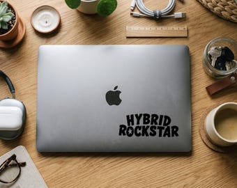 Op de afbeelding: Een zilveren laptop met een zwart Apple-logo en een zwarte sticker met de tekst "HYBRID ROCKSTAR". De laptop staat op een houten bureau, omringd door kantoorbenodigdheden, een kaars en een kleine potplant.