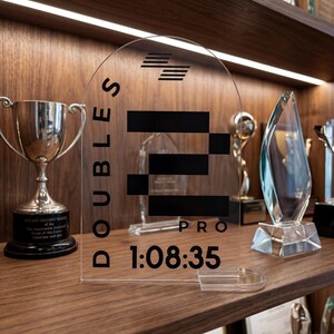 May include: A clear acrylic award with the text "DOUBLES PRO" and the time "1:08:35" printed in black. The award is displayed on a wooden shelf with other trophies and awards.