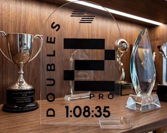Op de afbeelding: Een heldere acryl prijs met de tekst "DOUBLES PRO" en de tijd "1:08:35" in zwart gedrukt. De prijs wordt tentoongesteld op een houten plank met andere trofeeën en prijzen.