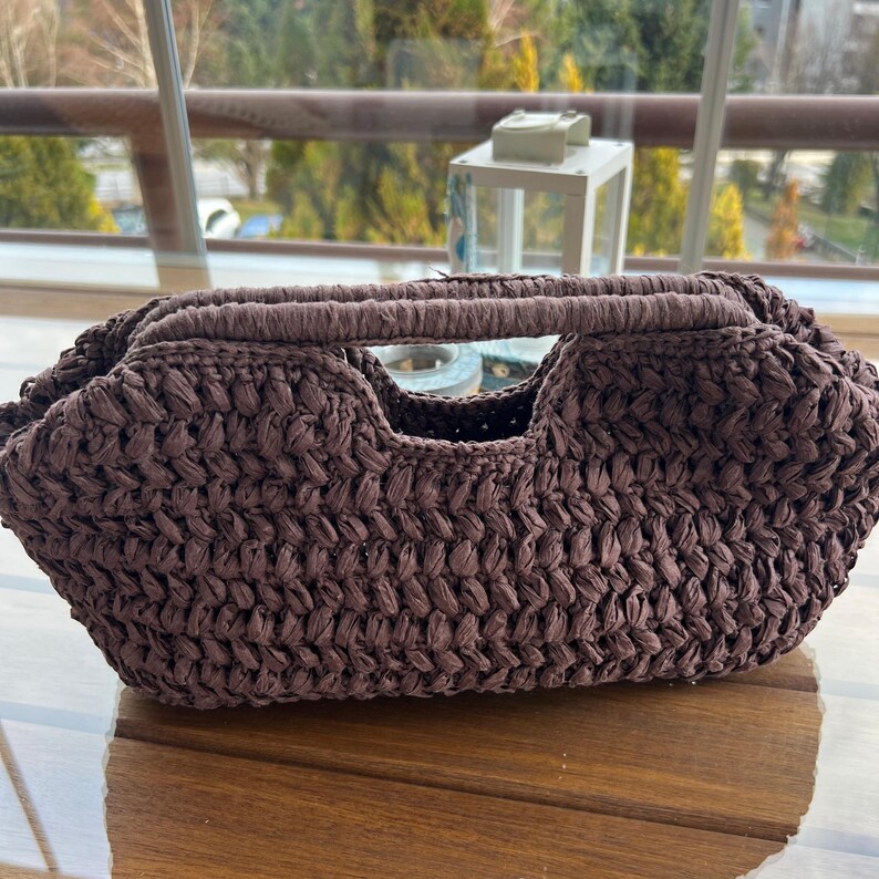 May include: A brown woven handbag with a handle and a rectangular shape. The bag has a textured surface and a cutout in the centre. The bag is on a wooden surface.