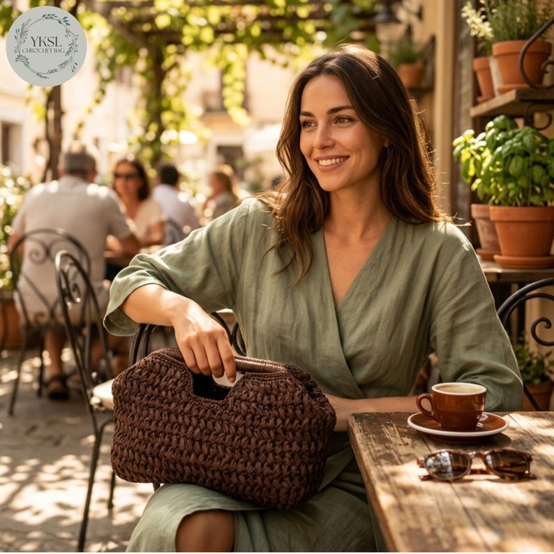 May include: A woman at a cafe table holds a brown woven handbag. She wears a green dress and smiles. A cup of coffee and sunglasses are on the wooden table. The bag has a distinctive handle. The image includes the text "YKSI CROCHETING".