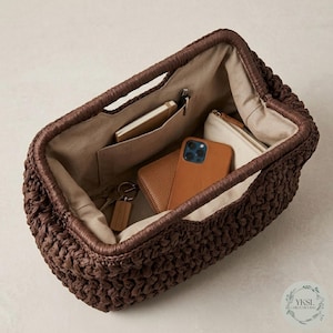 May include: A dark brown woven handbag with a rectangular shape, featuring a structured interior with pockets. Inside, a brown leather wallet, a smartphone, a small notebook, and a keychain are visible. The bag has a handle and a light-coloured interior lining.