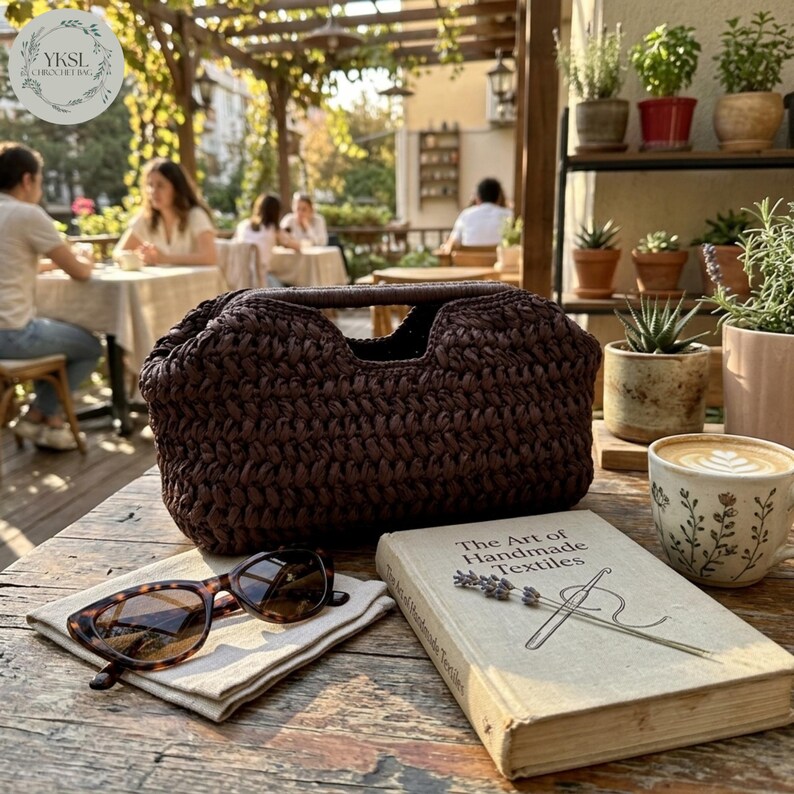May include: A dark brown crocheted handbag with a handle, on a wooden table. Tortoiseshell sunglasses sit on a white cloth next to a book titled "The Art of Handmade Textiles". A cup of coffee and potted plants are also visible.