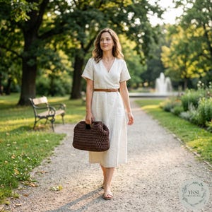May include: A woman walking in a park, carrying a dark brown crocheted handbag. She wears a cream-coloured wrap dress with a brown belt. The background includes trees, a bench, and a fountain.