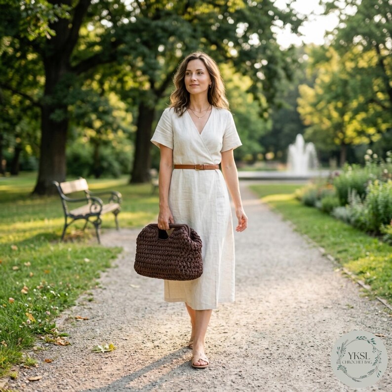 May include: A woman walking in a park, carrying a dark brown crocheted handbag. She wears a cream-coloured wrap dress with a brown belt. The background includes trees, a bench, and a fountain.