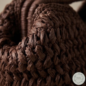 May include: Close-up of a woven brown bag. The bag is made of tightly woven, textured material, with a braided handle. The dark brown colour and intricate weave suggest a handmade or artisanal product, possibly a tote or basket.