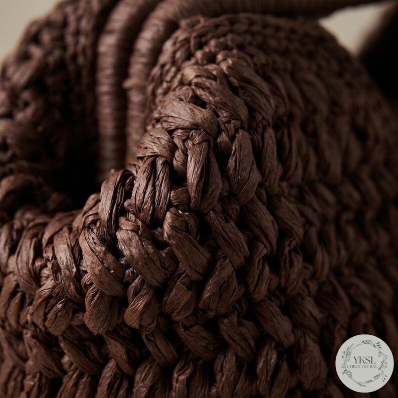 May include: Close-up of a woven brown bag. The bag is made of tightly woven, textured material, with a braided handle. The dark brown colour and intricate weave suggest a handmade or artisanal product, possibly a tote or basket.
