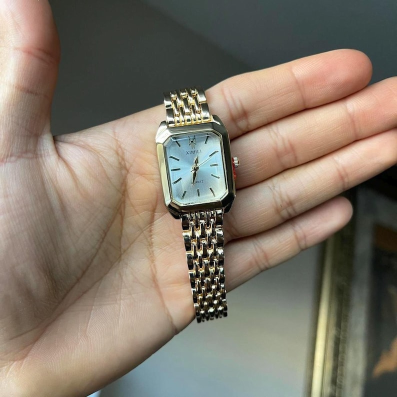 Op de afbeelding: Een rechthoekig horloge in goudtinten met een lichtgekleurde wijzerplaat en bijpassende band. De wijzerplaat heeft zwarte markeringen en wijzers, met de merknaam "XIMILI" zichtbaar. De band is een schakelontwerp.