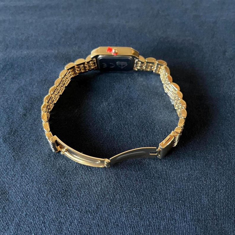 Op de afbeelding: Een goudkleurig polshorloge met een rechthoekige wijzerplaat en een schakelarmband. De wijzerplaat heeft een klein rood accent. Het horloge wordt open getoond, met de sluiting en de onderkant van de wijzerplaat zichtbaar.