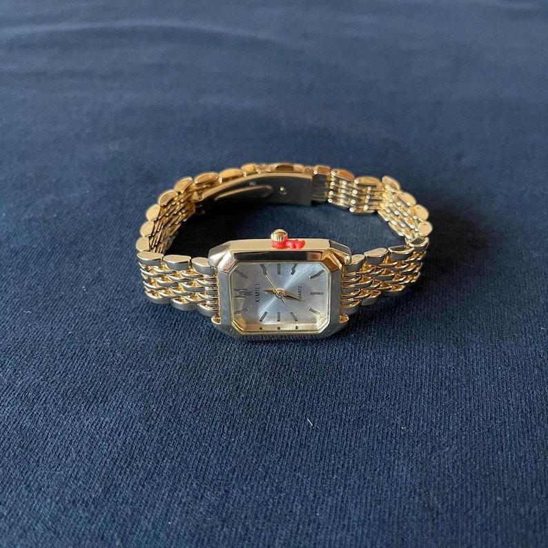 Op de afbeelding: Een goudkleurig polshorloge met een rechthoekige wijzerplaat en een schakelband. De wijzerplaat van het horloge is zilverkleurig met zwarte cijfers en wijzers. Het horloge staat tegen een donkerblauwe achtergrond.