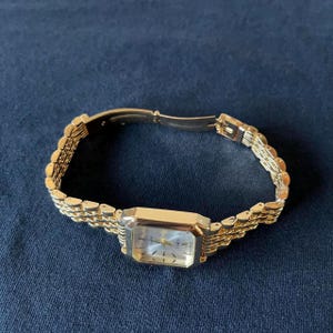 Op de afbeelding: Een goudkleurig polshorloge met een rechthoekige wijzerplaat en een schakelarmband. De wijzerplaat van het horloge is licht van kleur met gouden wijzers en markeringen. Het horloge ligt op een donkerblauwe ondergrond.