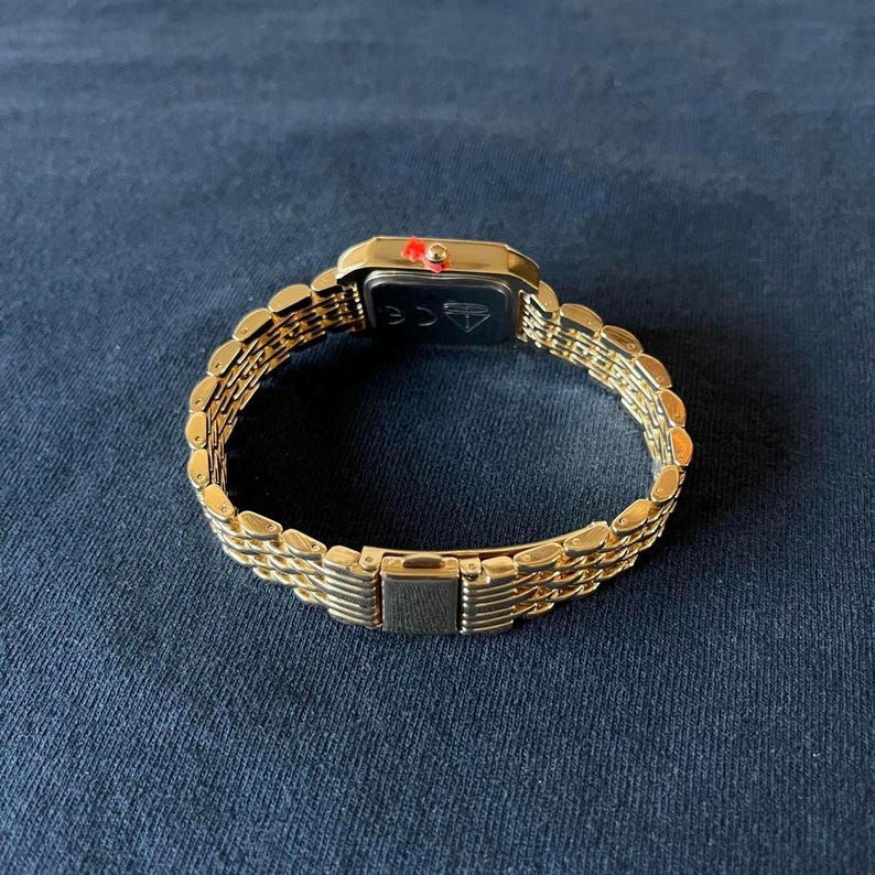 Op de afbeelding: Goudkleurig polshorloge met een rechthoekige zwarte wijzerplaat en een rode accent. Het horloge heeft een gouden schakelband en een sluiting. Het horloge wordt getoond op een donkerblauwe ondergrond.