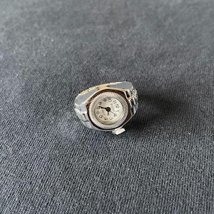 Puede incluir: Un reloj de anillo plateado con esfera blanca y números negros. El reloj tiene forma rectangular con una pequeña corona y un diseño tipo brazalete. El anillo está sobre un fondo de tela gris oscuro.