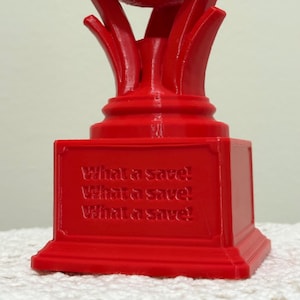 May include: A bright red trophy featuring a soccer ball resting on a stylized base. The square pedestal base has the text "What a save!" embossed three times in a stacked format on the front face, with a smooth, solid-colored plastic finish.
