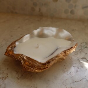 May include: A white candle in a gold-edged oyster shell. The candle has a single wick and is set on a marble surface. The oyster shell has a natural, textured appearance.