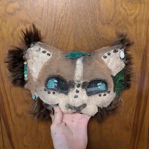 May include: A handmade lion mask with brown fur and a tan face. The mask features teal eyes, decorative beads, and green leaf accents. The mask is held up by a hand against a wooden door.