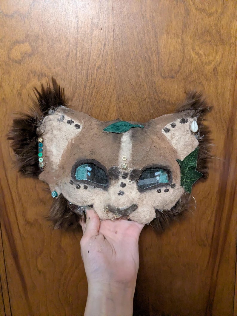 May include: A handmade lion mask with brown fur and a tan face. The mask features teal eyes, decorative beads, and green leaf accents. The mask is held up by a hand against a wooden door.