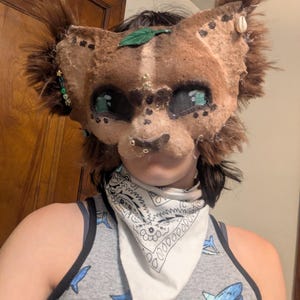 May include: A handmade, brown and tan feline mask with green and gold accents. The mask features large, expressive eyes, faux fur, and decorative embellishments. A green leaf and small shell adorn the mask. The person is wearing a gray tank top with blue shark graphics.