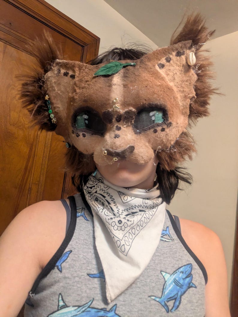 May include: A handmade, brown and tan feline mask with green and gold accents. The mask features large, expressive eyes, faux fur, and decorative embellishments. A green leaf and small shell adorn the mask. The person is wearing a gray tank top with blue shark graphics.