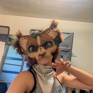 May include: A brown and tan animal mask with green accents and faux fur details. The mask has large, green eyes and is decorated with small gold-colored embellishments. The mask is designed to resemble a lion or similar feline.