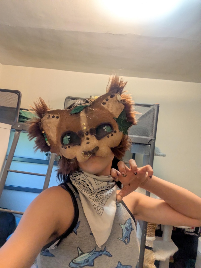 May include: A brown and tan animal mask with green accents and faux fur details. The mask has large, green eyes and is decorated with small gold-colored embellishments. The mask is designed to resemble a lion or similar feline.